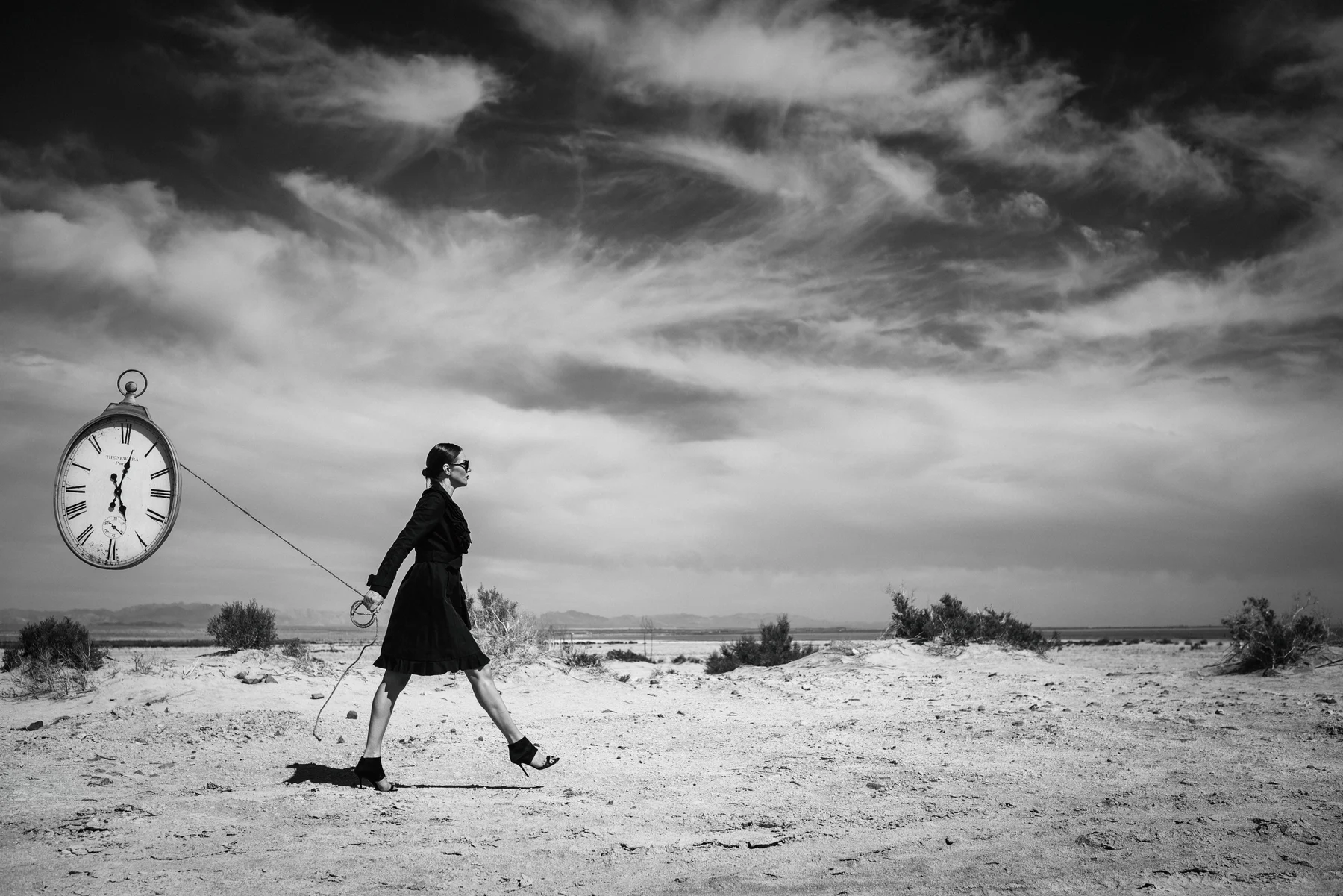 Temporal Baggage — An oversized clock follows a figure across the dry landscape. Photograph by Albert Pocej, California.
