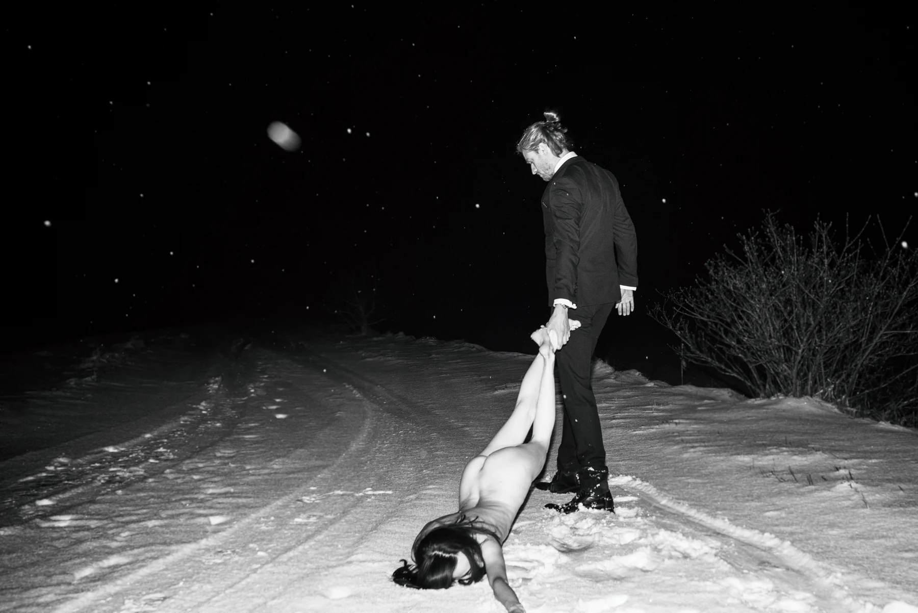 The Pull Of Love — A figure is held by the ankles on a snowy path at night. Photograph by Albert Pocej, Attachment Issues.