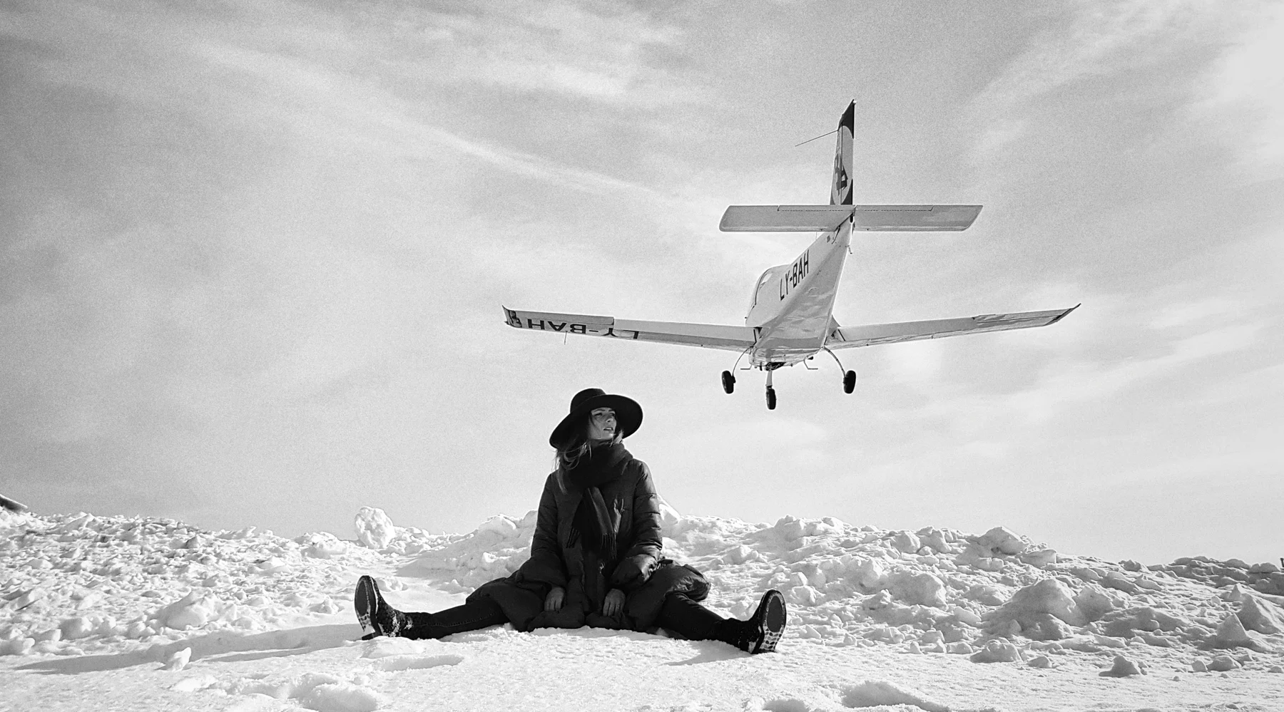 Waiting For Luggage — A woman sits in the snow beneath a low-flying aircraft. Photograph by Albert Pocej, Attachment Issues.