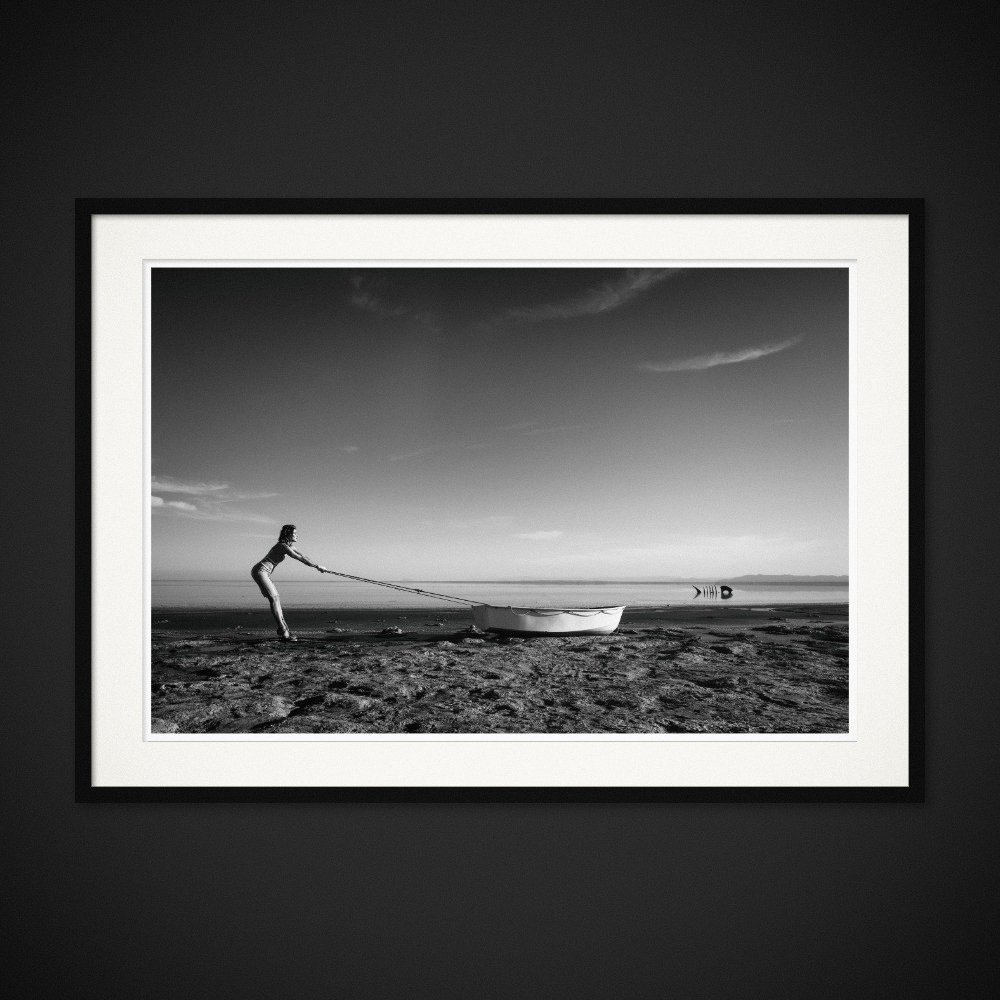 Echoes Of Rain — A small boat is pulled along the shore, opposite a fish-shaped structure. Framed edition by Albert Pocej.