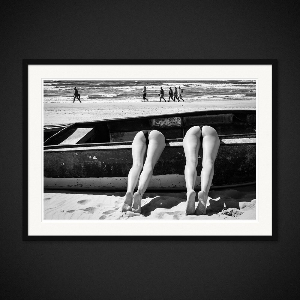 Sunbathers Anonymous — Two pairs of legs emerge from a boat on a busy beach. Framed edition by Albert Pocej.