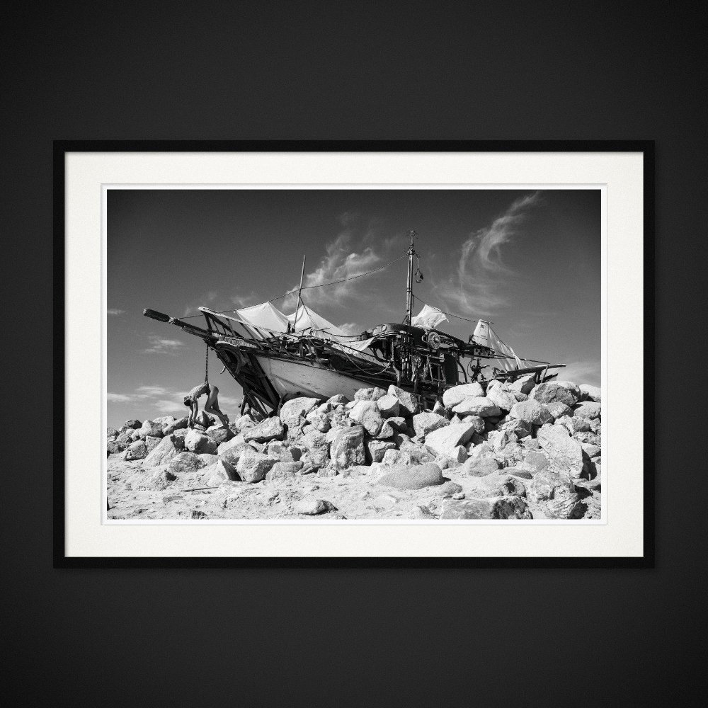 The Last Mermaid — A figure lies across a wrecked boat on a rocky shore. Framed edition by Albert Pocej.