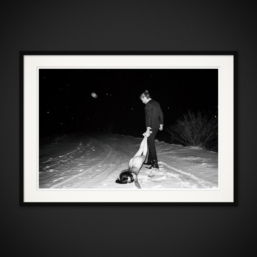 The Pull Of Love — A figure is held by the ankles on a snowy path at night. Framed edition by Albert Pocej.