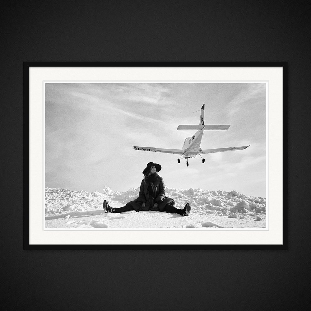 Waiting For Luggage — A woman sits in the snow beneath a low-flying aircraft. Framed edition by Albert Pocej.